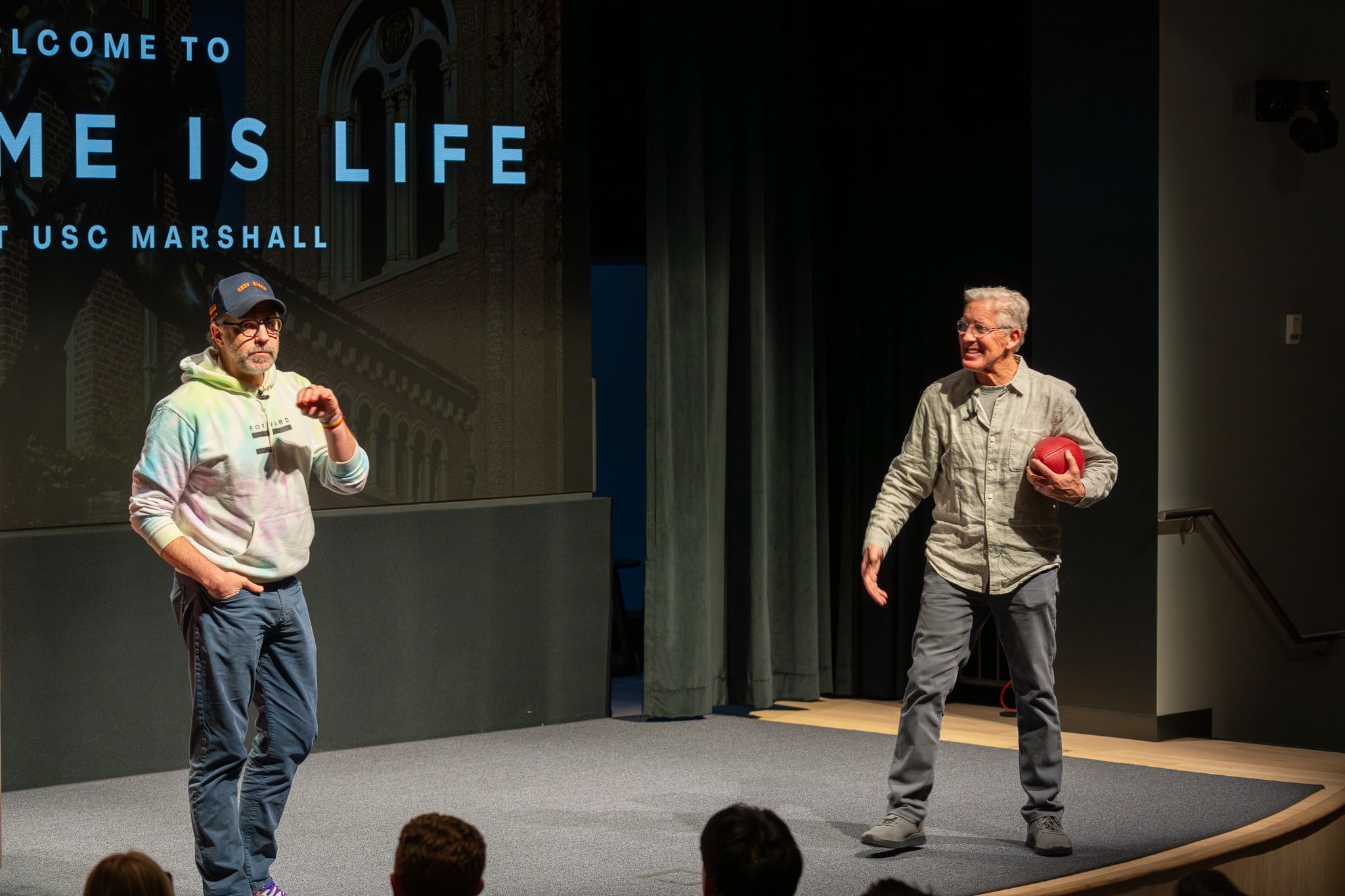 Pete Carroll catching a football tossed by Sudeikis in front of the Game is Life banner